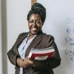 Smiling female lecturer holding books in front of classroom whiteboard.
